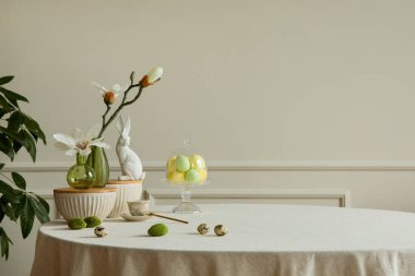 Minimalist composition of easter dining room interior with copy space, gray easter bunny sculpture, colorful eggs, vase with magnolia, beige wall with stucco and personal accessories. Home decor. Template.