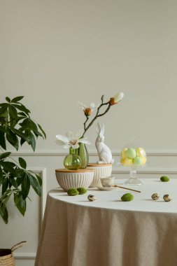 Interior design of dining room interior with round table, stylish beige bowl, colorful eggs, green glass vase with magnolia, white hare sculpture, cup and personal accessories. Home decor. Template.