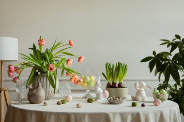 Interior design of easter dining room with colorful easter eggs, white hare sculptures, vase with tulips, plants, lamp, beige wall with stucco, gray hen and personal accessories. Home decor. Template.