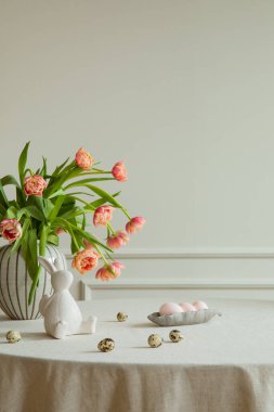 Minimalist composition of easter dining room interior with copy space, vase with tulips, colorful eggs, gray hen sculpture, beige wall with stucco and personal accessories. Home decor. Template.
