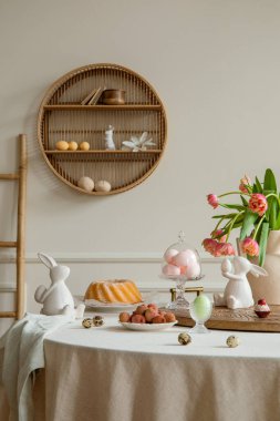 Interior design of easter dining room interior with round table, shelf on wall, colorful eggs, easter cake, hare sculptures, vase with tulips, lychee and personal accessories. Home decor. Template.