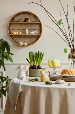 Warm and cozy composition of easter dining room interior with round table, cake, hare sculpture, colorful eggs, shelf on wall, wooden trace, plant and personal accessories. Home decor. Template.