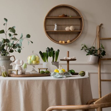 Warm and spring dining room interior with easter accessories, shelf on wall, round table, vase with green leaves, colorful eggs, rabbit sculpture and personal accessories. Home decor. Template.