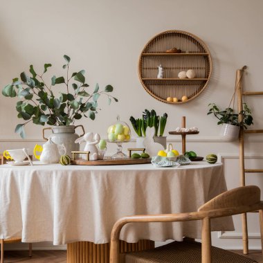 Warm and spring dining room interior with easter accessories, round table, vase with green leaves, cake, colorful eggs, rabbit sculpture and personal accessories. Home decor. Template.