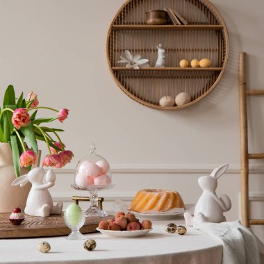 Interior design of easter dining room interior with round table, shelf on wall, colorful eggs, easter cake, hare sculptures, vase with tulips, lychee and personal accessories. Home decor. Template.