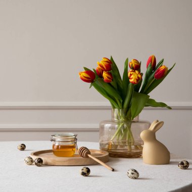 Minimalist composition of easter dining room interior with copy space, round table, vase with tulips, easter bunny sculpture, honey in jar, beige wall and personal accessories. Home decor. Template.