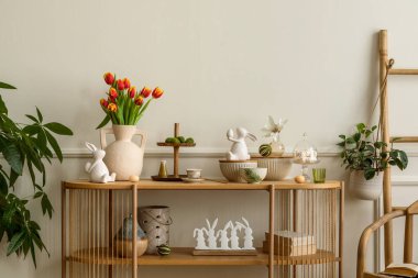 Warm and cozy composition of easter  living room interior with mock up poster frame, vase with tulips, hare sculpture, carrot, bowl, wooden sideboard and personal accessories. Home decor. Template.