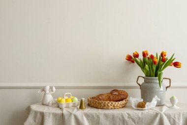 Minimalist composition of living room nterior with copy space, vase with tulips, bread in basket, colorful easter eggs, easter bunny sculpture and personal accessories. Home decor. Template.
