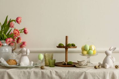 Creative composition of easter living room interior with copy space, vase with spring tulips, easter bunny sculpture, elegant cup, colorful easter eggs and personal accessories. Home decor. Template.