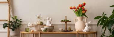 Warm and cozy composition of easter  living room interior with mock up poster frame, vase with tulips, hare sculpture, carrot, bowl, wooden sideboard and personal accessories. Home decor. Template.