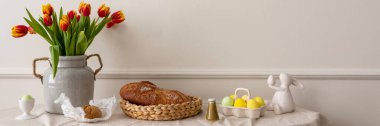 Minimalist composition of living room nterior with copy space, vase with tulips, bread in basket, colorful easter eggs, easter bunny sculpture and personal accessories. Home decor. Template.