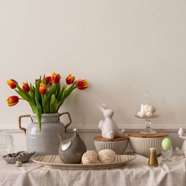 Minimalist composition of living room nterior with copy space, vase with tulips, bread in basket, colorful easter eggs, easter bunny sculpture and personal accessories. Home decor. Template.