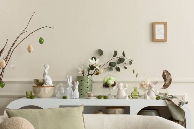Interior design of easter living room interior with mock up poster frame, stylish sideboard, colorful easter eggs, easter bunny sculpture, vase with leaves and personal accessories Home decor Template