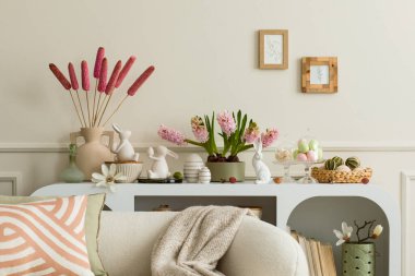 Interior design of easter living room interior with mock up poster frame, stylish white sideboard, hyacinth, easter bunny, vase with dried flowers and personal accessories. Home decor. Template.