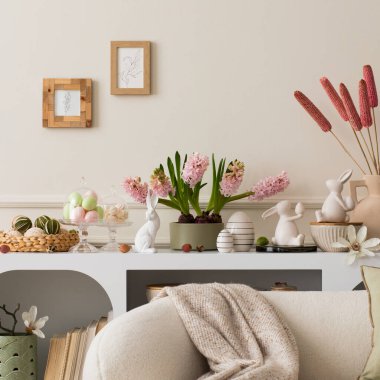Interior design of easter living room interior with mock up poster frame, stylish white sideboard, hyacinth, easter bunny, vase with dried flowers and personal accessories. Home decor. Template.