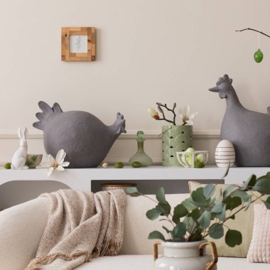 Creative composition of easter living room interior with mock up poster frame, hen sculpture, easter eggs, easter bunny, white sofa, beige pillows and personal accessories. Home decor. Template.