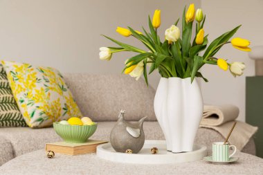 Tarafsız renk paleti olan Paskalya temalı bir oturma odası. Seramik tavşan heykelcikleri, pastel yumurtalar ve beyaz bir vazoda taze sarı lalelerle süslenmiş yuvarlak bir sedir, şenlikli bir dokunuş ekler..