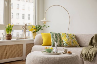 Tarafsız renk paleti olan Paskalya temalı bir oturma odası. Seramik tavşan heykelcikleri, pastel yumurtalar ve beyaz bir vazoda taze sarı lalelerle süslenmiş yuvarlak bir sedir, şenlikli bir dokunuş ekler..