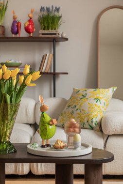 Tarafsız renk paleti olan Paskalya temalı bir oturma odası. Seramik tavşan heykelcikleri, pastel yumurtalar ve beyaz bir vazoda taze sarı lalelerle süslenmiş yuvarlak bir sedir, şenlikli bir dokunuş ekler..