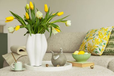 Sarı laleli Paskalya 'dan esinlenilmiş ev dekoru seramik bir tavuk ve eskimiş yeşil bir kâsede pastel renkli yumurtalar. Tarafsız bir arkaplan canlı mevsimsel detayları geliştirir.