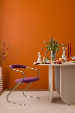 Pastel süslemeler, laleler, seramik tavşanlar ve sıcak turuncu bir duvara dayalı ahşap bir masa..