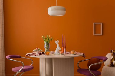 Pastel süslemeler, laleler, seramik tavşanlar ve sıcak turuncu bir duvara dayalı ahşap bir masa..