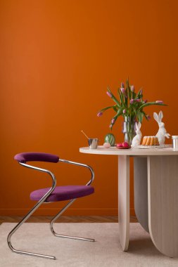 Pastel süslemeler, laleler, seramik tavşanlar ve sıcak turuncu bir duvara dayalı ahşap bir masa..