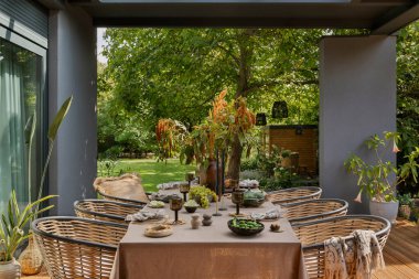 Masa, bahçe mobilyaları, çiçekli vazo, ahşap döşeme, ekose, perde, üzüm, büyük bahçe ve kişisel aksesuarlı yaratıcı kompozisyon. Ev dekoru. Şablon.