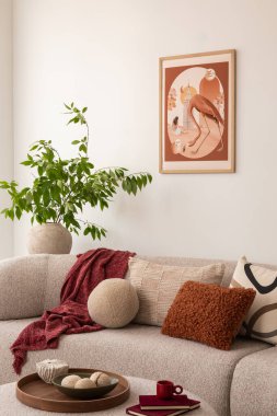 Oturma odasında bej bir kanepe ve desenli yastıklarla süslenmiş. Duvarda bir kuşla bir kadını gösteren çerçeveli bir sanat eseri asılı. Saksı bitkisi ve açık bir kitap dingin atmosfere katkı sağlar..