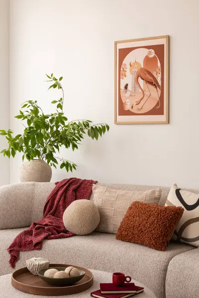 Oturma odasında bej bir kanepe ve desenli yastıklarla süslenmiş. Duvarda bir kuşla bir kadını gösteren çerçeveli bir sanat eseri asılı. Saksı bitkisi ve açık bir kitap dingin atmosfere katkı sağlar..