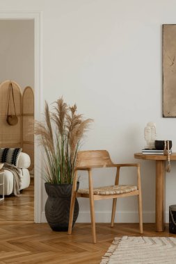 Minimalist bir tasarımı olan modern bir oturma odası. Dekoratif vazolar ve heykellerle süslenmiş ahşap bir masa, örülmüş koltuklu ahşap bir sandalye ve duvarda büyük soyut bir tablo yer alıyor. Doğal ışık, sakin ve rahat atmosp 'u vurgular