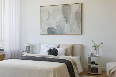 Yumuşak nötr tonları olan parlak minimalist yatak odası yatağın üzerinde büyük soyut resim ve beyaz perdelerin arasından akan doğal ışık..