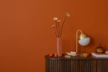 Dekoratif nesnelerle donatılmış minimalist bir iç mekan ve ahşap bir dolapta pembe bir tırtıklı vazo, yanmış turuncu bir duvara karşı. Sakin, modern kompozisyon. Boşluğu kopyala.
