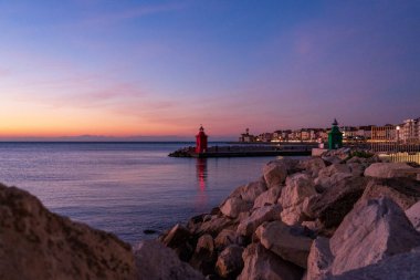 Piran, Slovenya - 3 Kasım 2024: Denize karşı kırmızı deniz feneri. Liman şehrinde akşam batımı. Suda kayalar olan deniz feneri. Piranski svetilnik