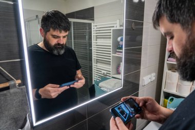 Aynada sakallı bir adam akıllı telefonuna bakar. Banyo..