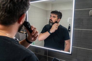 Adam banyoda sakalını kesiyor. Sakal bakımı, elektrikli jilet. Banyo aynasında sakallı bir adamın yansıması..