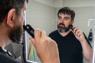 Adam banyoda sakalını kesiyor. Sakal bakımı, elektrikli jilet. Banyo aynasında sakallı bir adamın yansıması..
