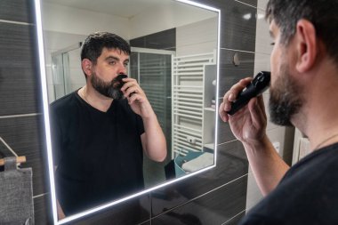 Adam banyoda sakalını kesiyor. Sakal bakımı, elektrikli jilet. Banyo aynasında sakallı bir adamın yansıması..