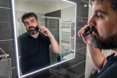 Adam banyoda sakalını kesiyor. Sakal bakımı, elektrikli jilet. Banyo aynasında sakallı bir adamın yansıması..