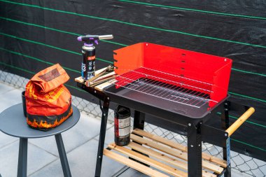 Sıcak bir açık verandada kırmızı bir mangal mangalı kurulur. Yakınlarda gerekli ızgara aletleri ve gaz lambası, çakmak gazı ve tütsü gibi kullanıma hazır eşyalar bulunuyor. Bu görüntü, barbekü hazırlama sahnesini rahat ve durgun bir şekilde yakalar.