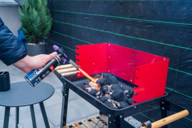 A man's hand with a gas torch lights briquettes on a portable red grill on an outdoor patio. Nearby are the necessary grilling tools and items, such as a gas torch and lighter fluid, ready to use. This image captures the preparation stage of a barbec clipart