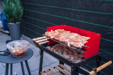 A vibrant red barbecue grill loaded with marinated meat, sizzling over hot charcoal briquettes in an outdoor patio setting. The scene is illuminated for evening cooking, surrounded by cozy decor and evergreen plants. This image is perfect for illustr clipart