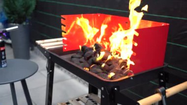 Sıcak bir açık verandada kırmızı bir mangal mangalı kurulur. Yakınlarda gerekli ızgara araçları ve gaz lambası, çakmak gazı, briketler gibi kullanıma hazır eşyalar bulunuyor..