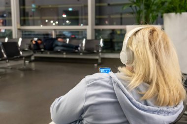 Viyana, Avusturya - 19 Ocak 2025: Havaalanı terminalinde kulaklık takan bir kadın yolcu. Uçağa binmeden önce uçak bekliyor. Bekleme odasındaki koltuklarda uyuyan bir adam..