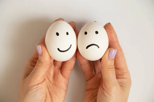 Two eggs with a joyful smile and a sad one in the hands of a girl on a white background, joy and sadness choice, lunch
