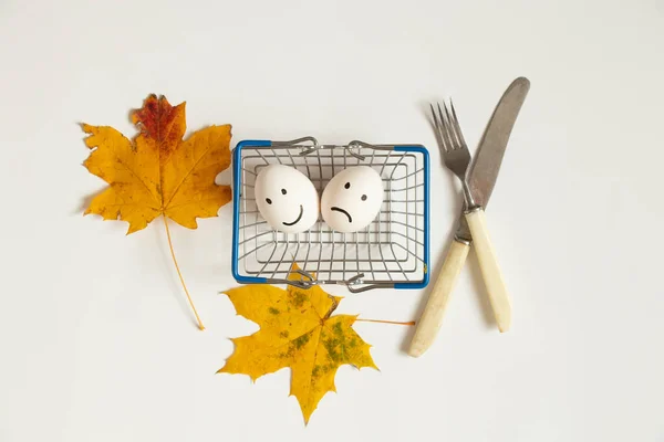 Two eggs with a joyful smile and a sad one lie in a supermarket basket next to autumn leaves and a knife with a fork on a white background, a choice of joy and sadness, lunch