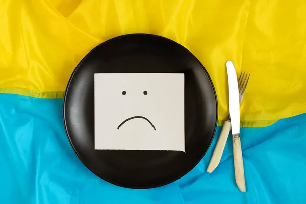 A sad emoticon drawn on paper lies in a black plate on the flags of Ukraine, a bad mood, no food and hunger due to the war 2022