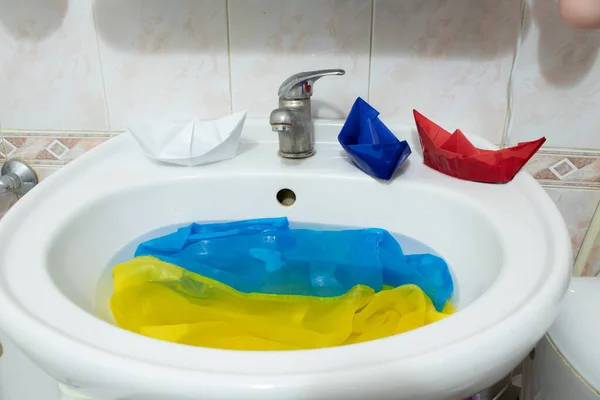 The flag of Russia from paper boats floats on the water on the flags of Ukraine in the sink in the bathroom, the war in Ukraine with Russia