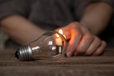 An incandescent light bulb and a candle in the hands of an old woman on a wooden table, people without light, the light of a candle when there is no light in Ukraine