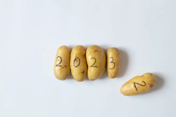 Four raw potatoes with the numbers 2023 and next to them lies a potato with the number 2, written in black felt-tip pen, lie on a white background, vegetables in the new year